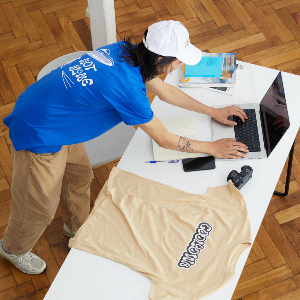Maximize sales during seasonal trends with Printify Man in a blue shirt and white cap types on a laptop at a white table, surrounded by a notebook, phone, camera, and beige t-shirt.