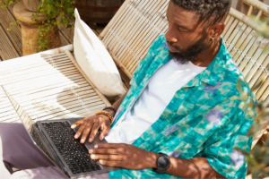 A man in a colorful shirt focuses on using a laptop while sitting on a bamboo bench outdoors.