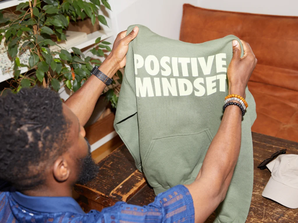 A man holds up an olive green hoodie with "POSITIVE MINDSET" printed in bold white text.