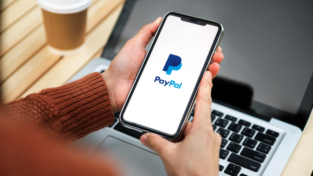 A person holds a smartphone displaying the PayPal logo, situated above a laptop keyboard.