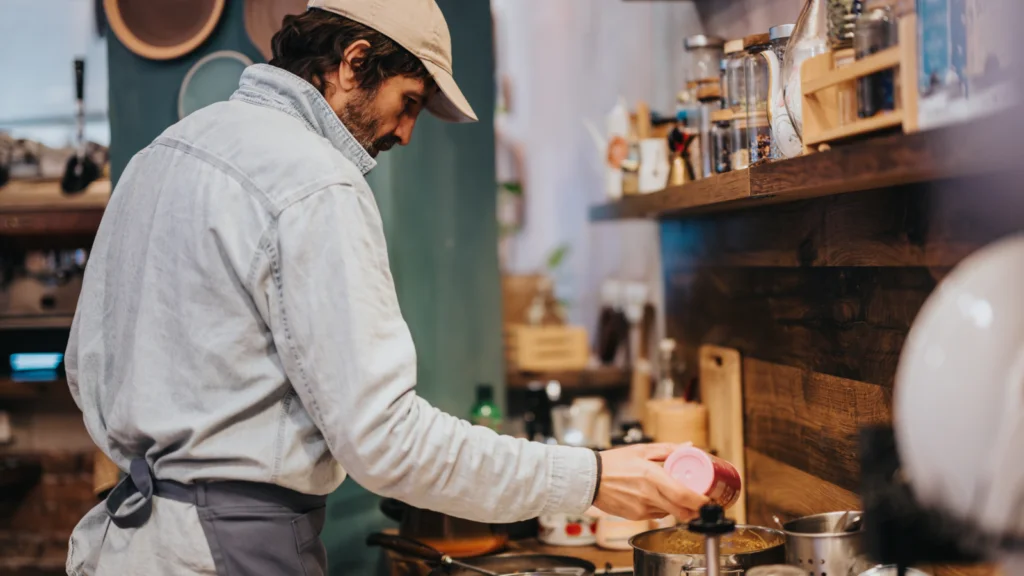 A man wearing a hat is preparing food in a kitchen, focused on his cooking tasks.