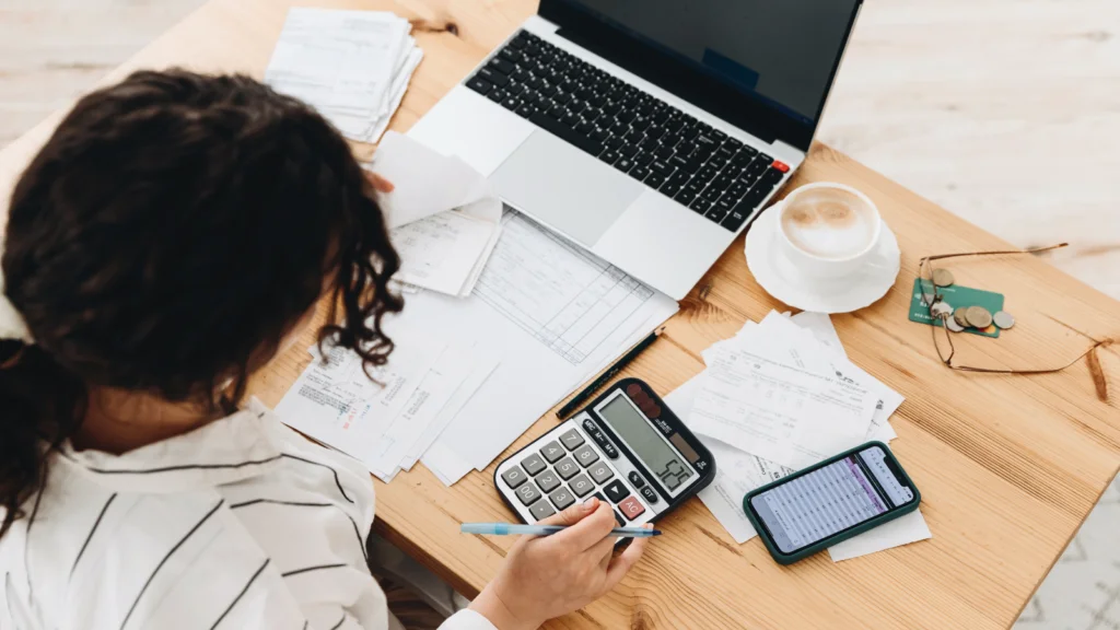 A woman sits at a wooden table with a laptop, calculator, documents, and a smartphone. A cup of coffee and glasses are nearby.
