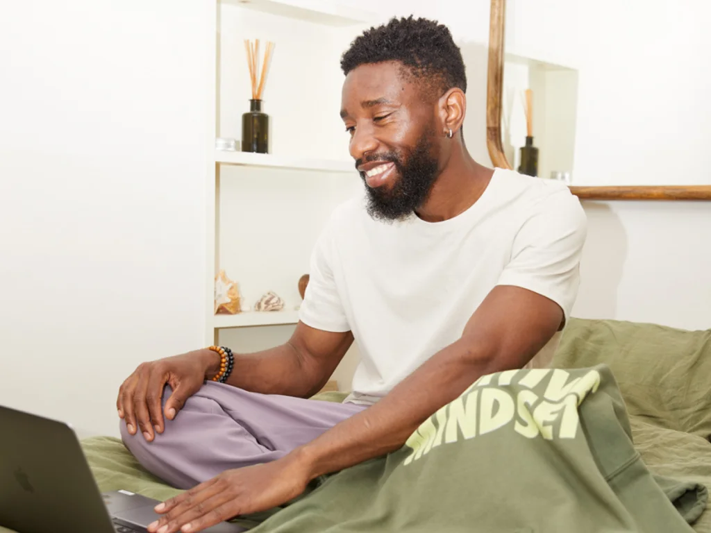 A man sits on a bed, smiling at a laptop, wearing a white shirt and purple pants.