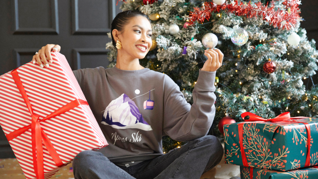 Seasonal gifts and holiday ready decor A woman in a gray sweater sits joyfully by a decorated Christmas tree holding a bauble. She is surrounded by red and green wrapped gifts.