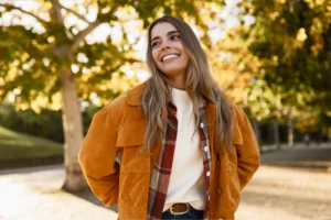 Smiling woman outdoors in autumn, wearing a brown jacket over a plaid shirt and white sweater. Trees with golden leaves in the backdrop.