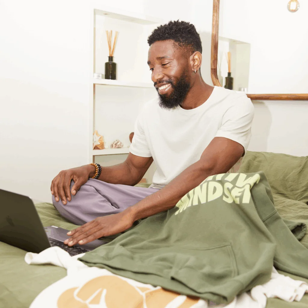 Sell unique print on demand products with Printify A smiling man is sitting on a bed with a laptop and a green hoodie with “Positive mindset” print nearby.