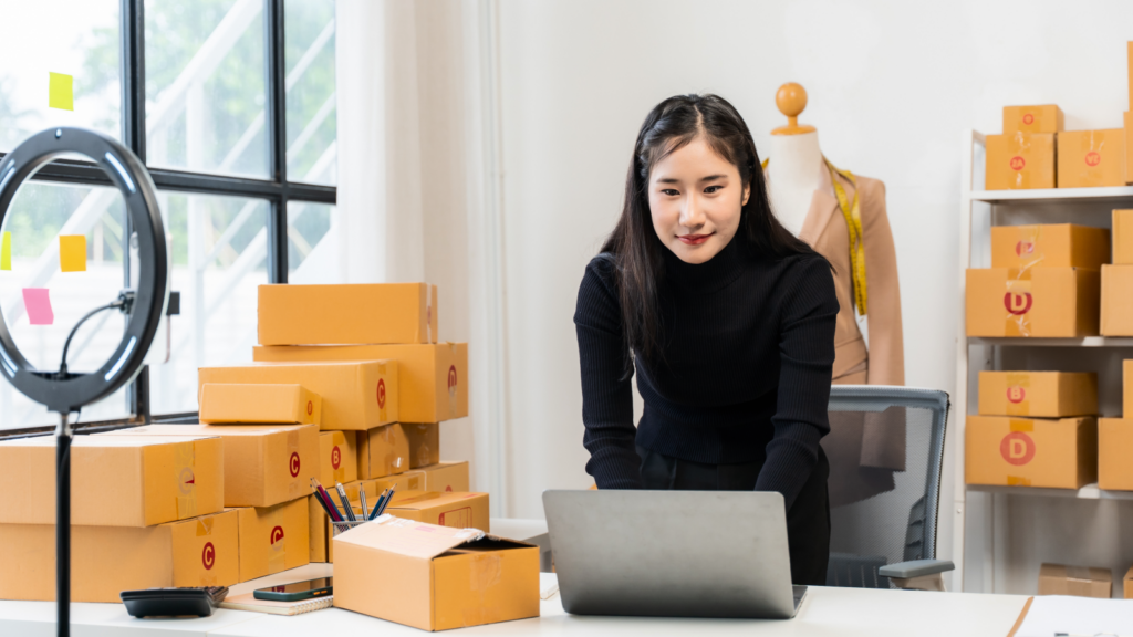 A woman in a black sweater works at a desk with a laptop, learning how to start a skincare line, surrounded by stacked cardboard boxes.