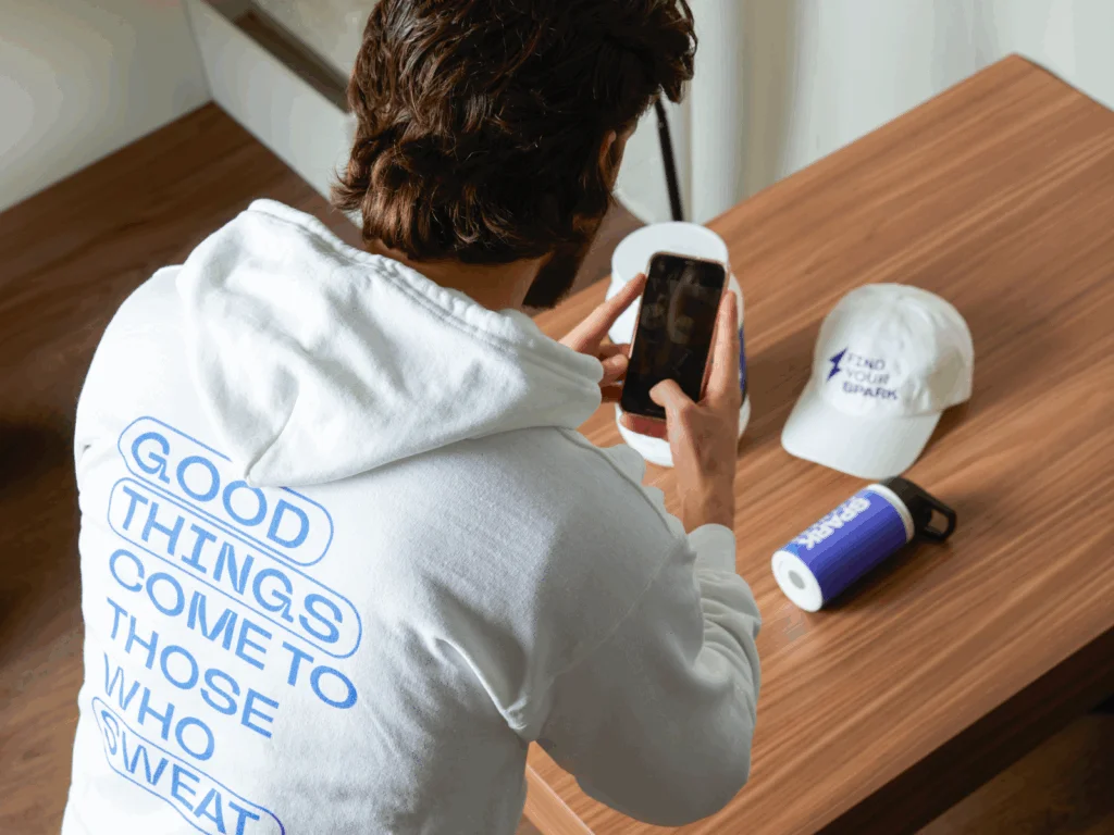 A man in a white hoodie with "Good Things Come To Those Who Sweat" photographs a water bottle and a hat.