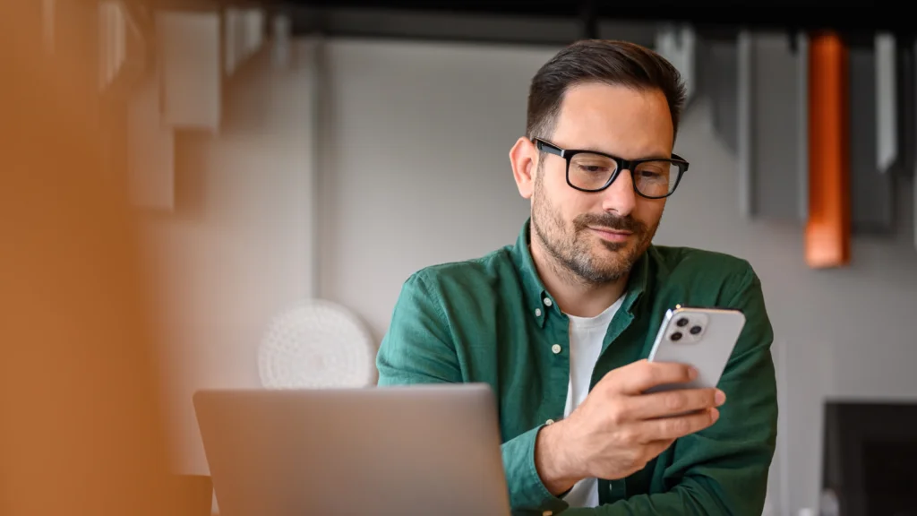 A man wearing glasses and a green shirt sits at a desk, looking at a smartphone as he searches for the best TikTok tools.