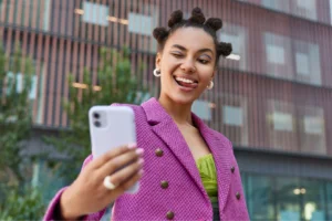 A young woman in a vibrant pink blazer winks playfully while taking a selfie outside a modern building.