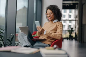 A woman with curly hair smiles while using a smartphone, seated in a bright room. She has a laptop on her lap, looking happy.