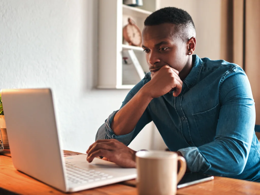 A man in a denim shirt focused on a laptop screen at a wooden desk, with a beige mug in the foreground.