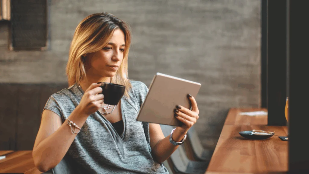 A woman in a gray shirt holds a tablet and a black coffee mug in a cafe, while searching for clothing brand name ideas.