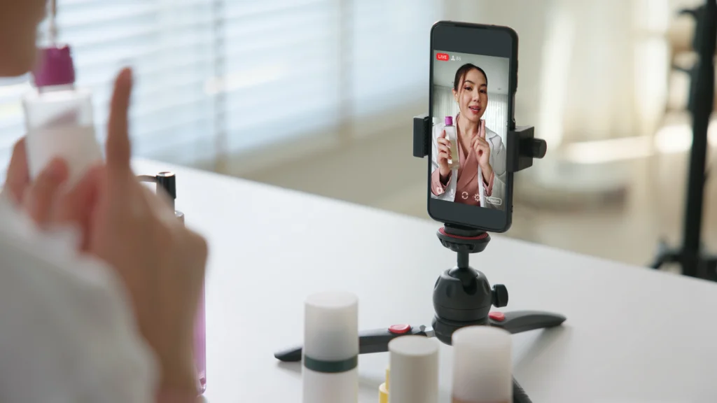 A woman livestreams, holding a skincare product, phone mounted on a tripod.