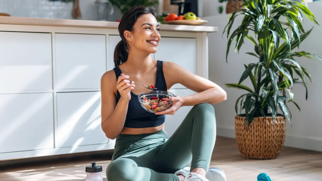 A woman in workout clothes sits on a kitchen floor, smiling while holding a bowl of fruit salad. Nearby are a water bottle and a potted plant.