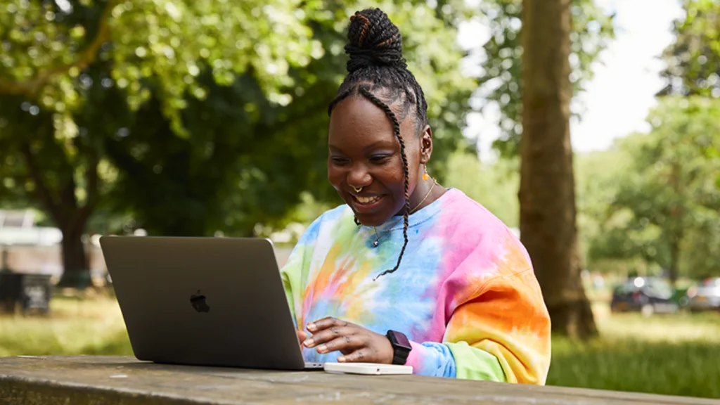 What does this mean for POD sellers A woman in a tie-dye sweatshirt smiles while searching how AI predicts viral designs on a laptop. Seated at a wooden bench in a park.