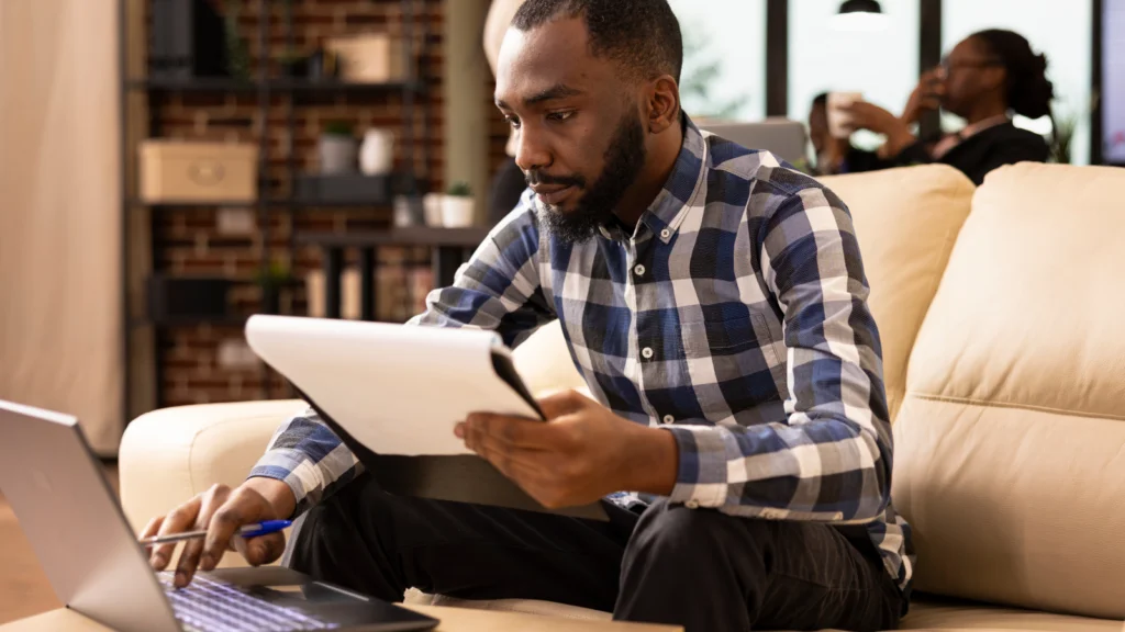 A man in a plaid shirt works on a laptop while holding documents. He sits on a beige sofa in a cozy, well-lit living room.
