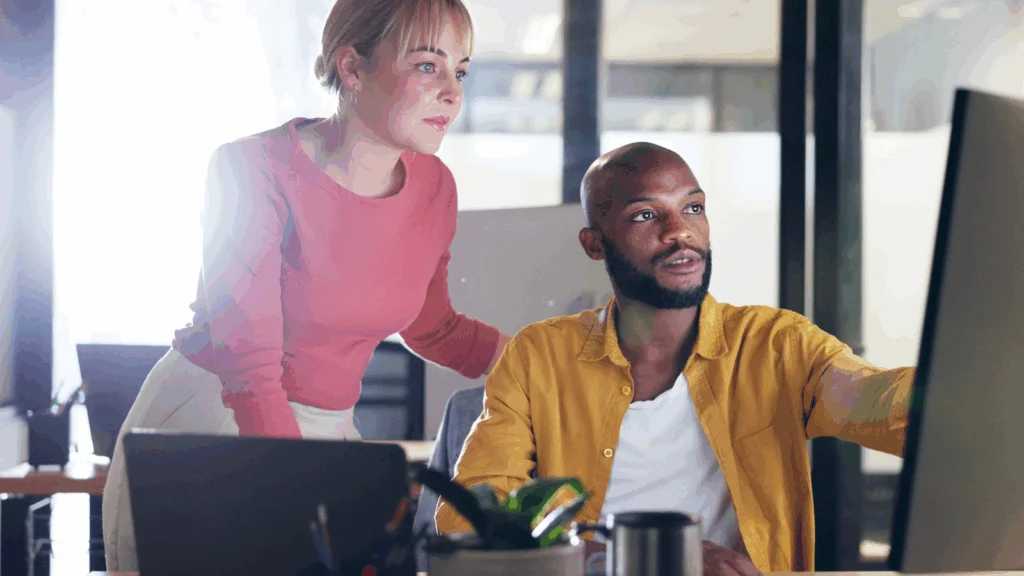 A woman in a pink sweater and a man in a yellow shirt collaborate in an office, focused on a computer screen.