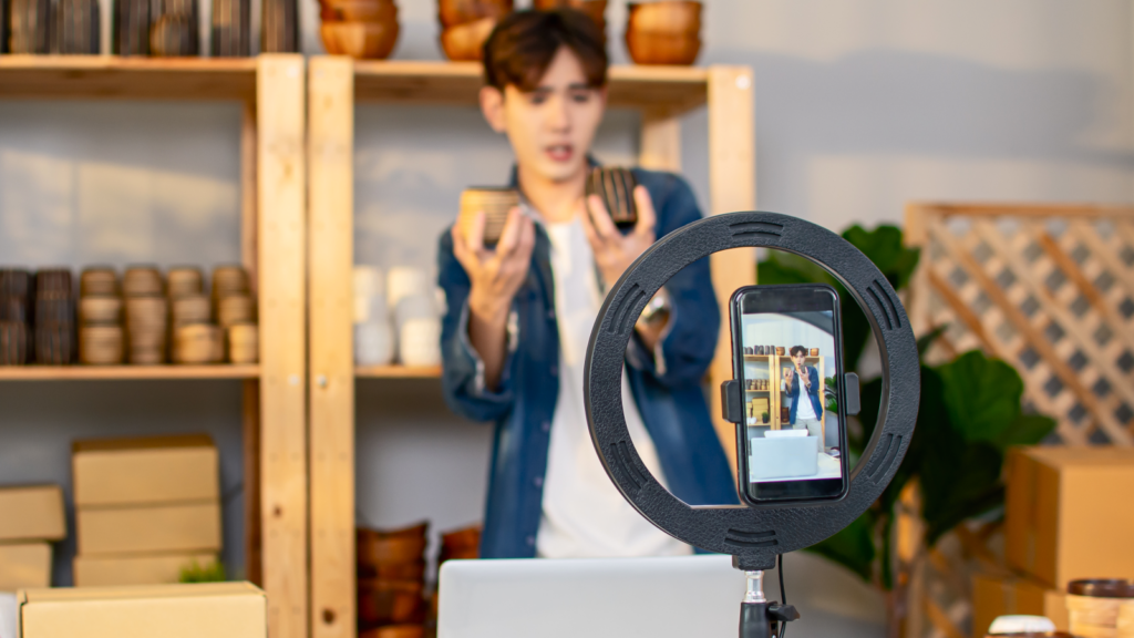 A person is livestreaming using a phone, holding two products while standing in front of wooden shelves with boxes.