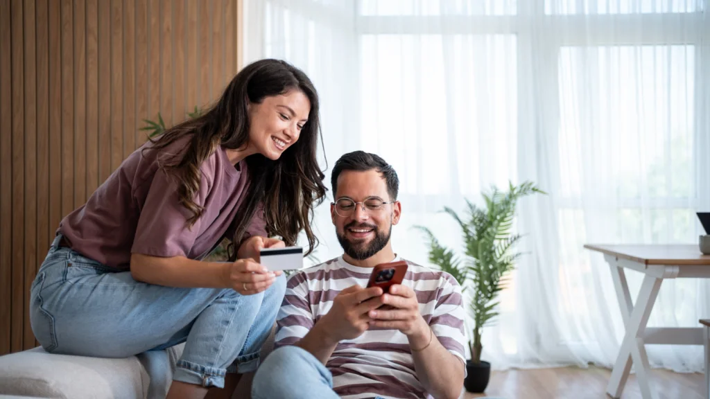 Smiling man and woman in casual clothes indoors, man holding a phone and woman holding a credit card.