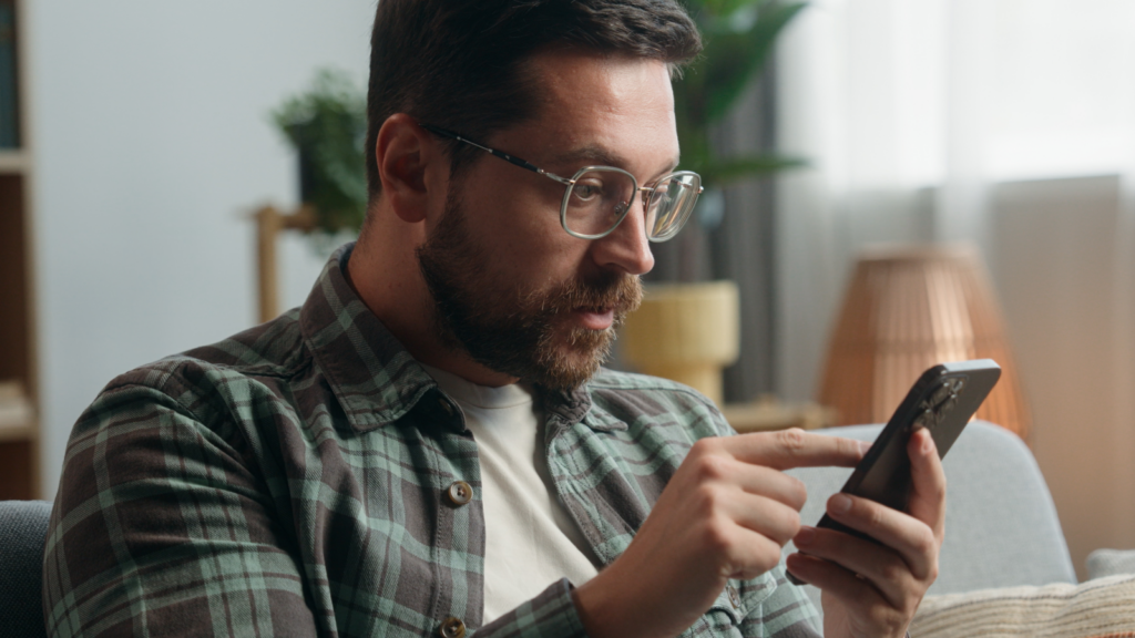 A man with glasses and a beard uses a smartphone on a couch to research TikTok Promotion. He wears a plaid shirt.