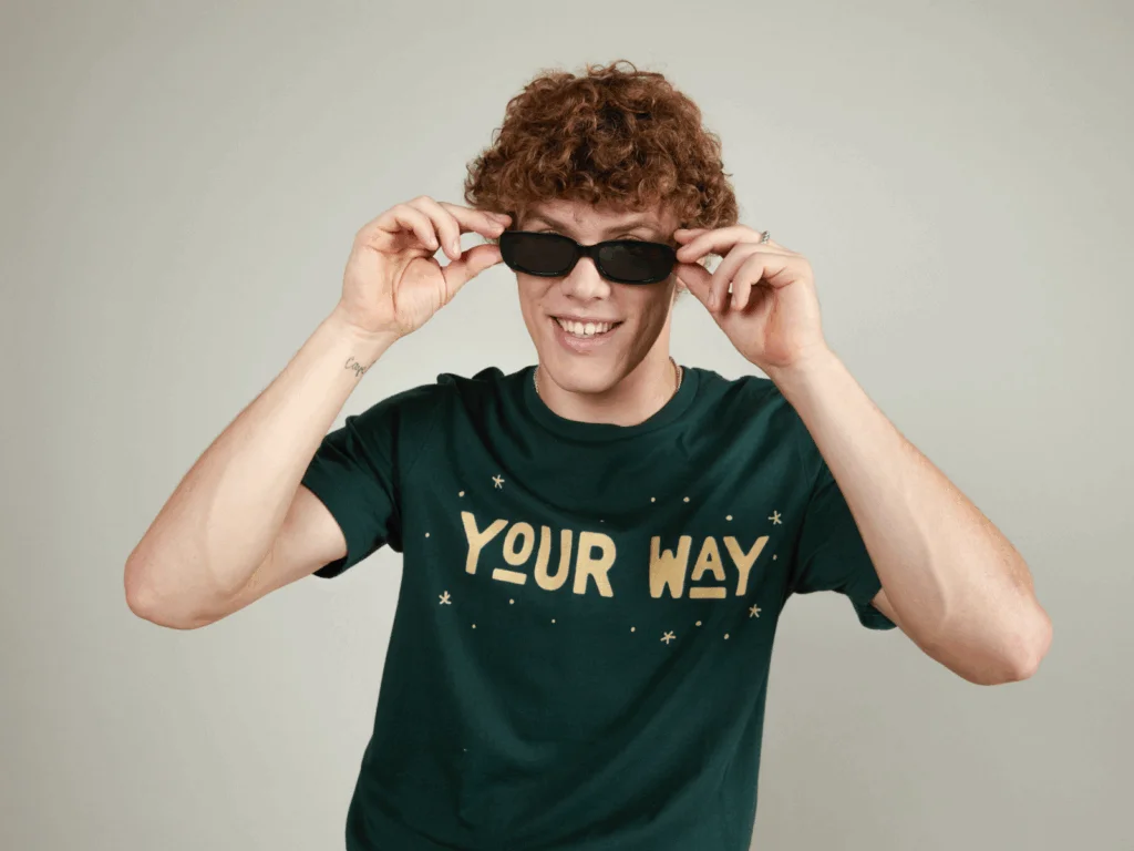 A young man with curly hair smiles and puts on sunglasses. He wears a dark green shirt with "Your Way" in bold letters.