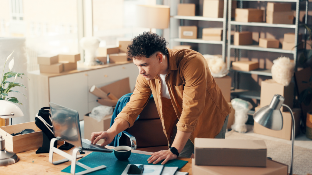 A man in a casual shirt works on a laptop, searching for “What is Klarna”, in a sunlit office filled with cardboard boxes.