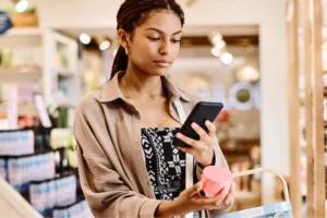 How to price a product Printify A woman holding a shopping bag while looking at her phone, appearing engaged in a conversation or shopping online.