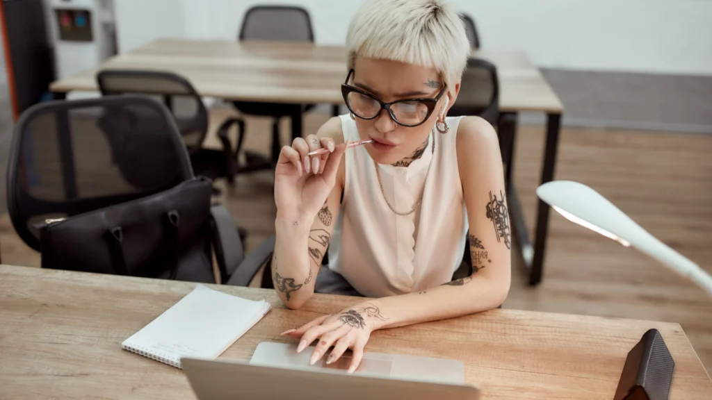 A woman with tattoos and short blond hair sitting in an office is using a laptop to find Redbubble alternatives, holding a pencil near her lips.