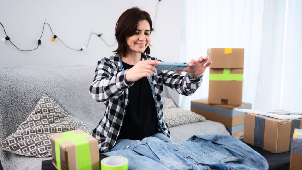 A smiling woman wearing a plaid shirt takes a photo of a denim jacket. Surrounding her are cardboard boxes, tape, and a sofa.