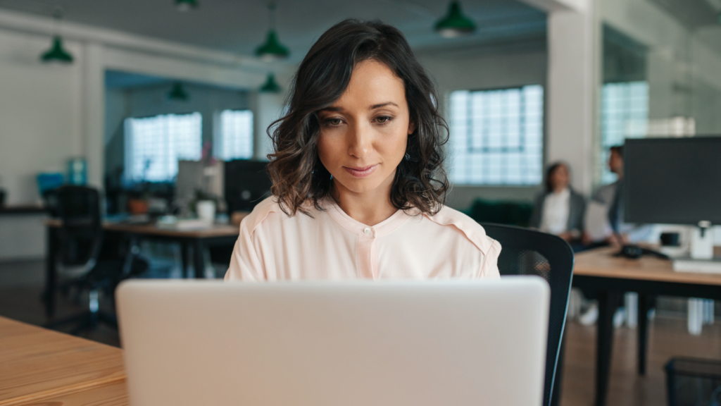 A woman with focuses on her laptop in a modern office setting. The background is softly blurred.