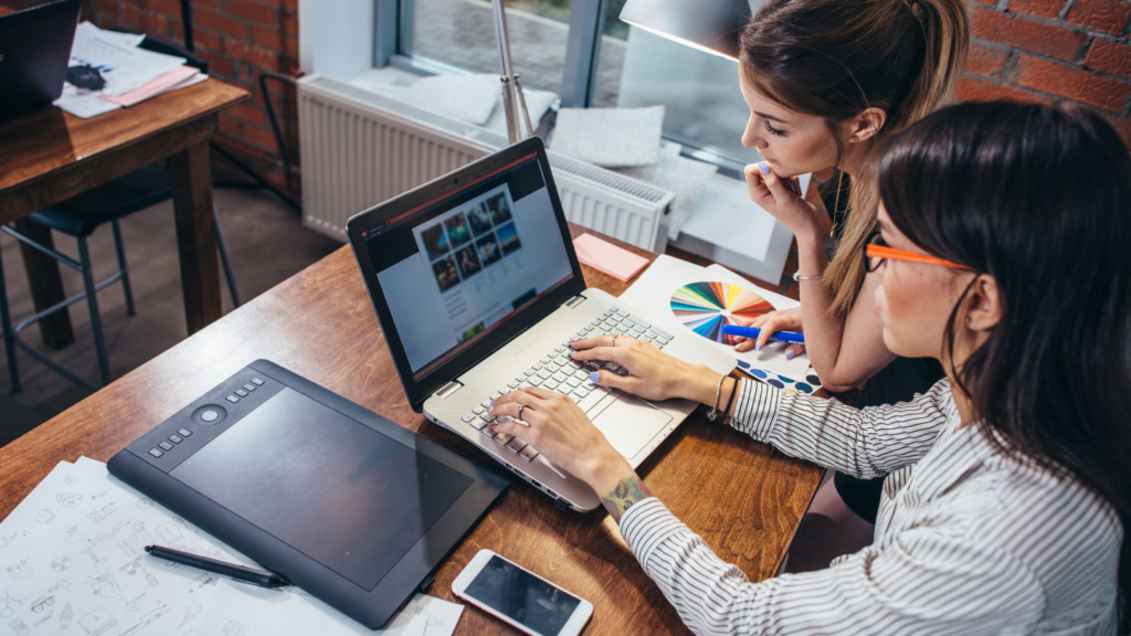 Two women work together on a laptop at a wooden desk, reviewing graphic designs, surrounded by design tools.