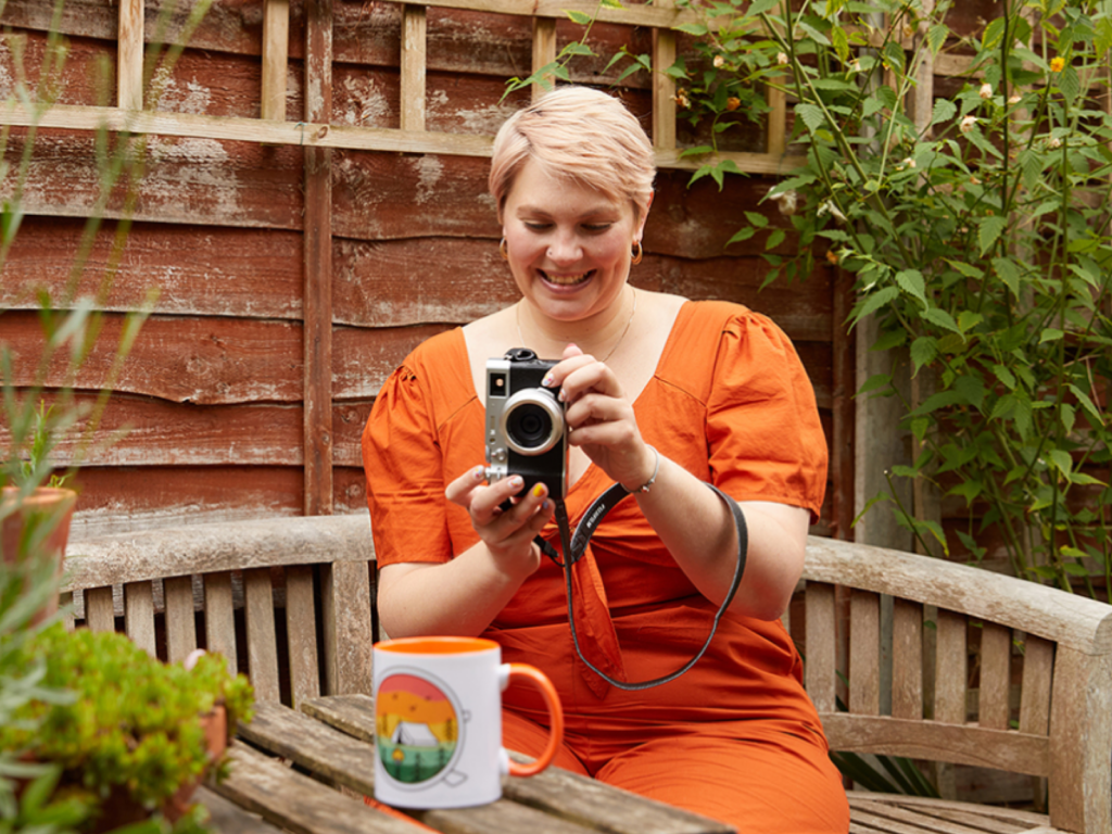 A smiling young woman is taking a picture of a mug with a DSLR camera in a small garden area.