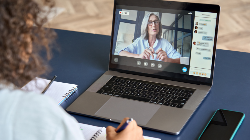 A person writing in a notebook while watching an online course on a laptop. The screen shows a woman teaching online.