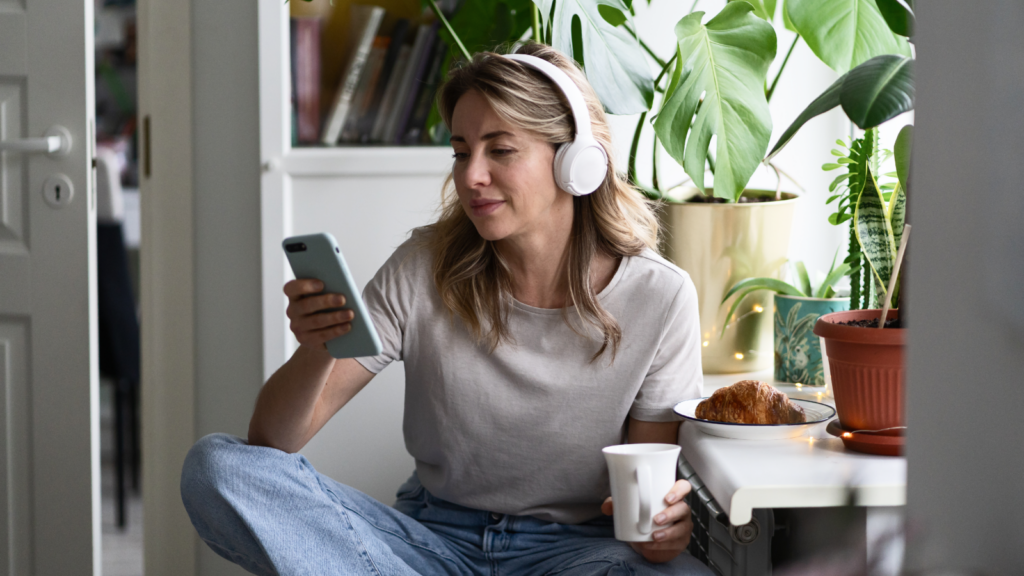 A woman sits comfortably in a kitchen, wearing headphones, looking at her phone. She's holding a mug, with plants and a croissant nearby.