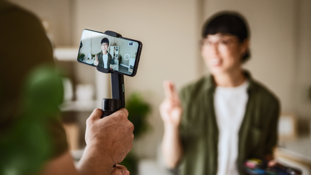 A person is filming a woman with a smartphone on a gimbal, smiling and gesturing with fingers crossed.