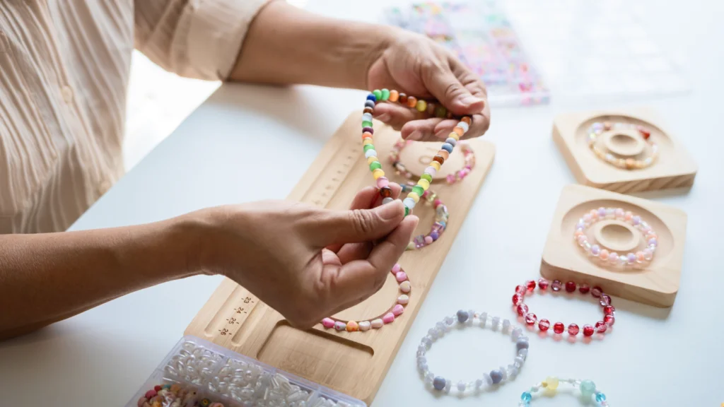 A person is crafting beaded bracelets on a wooden board. The table has colorful beads and completed bracelets.