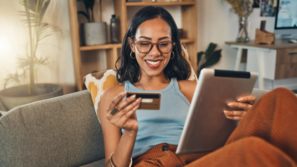 A woman with glasses holds a credit card while sitting on a couch in her home, looking relaxed and focused.