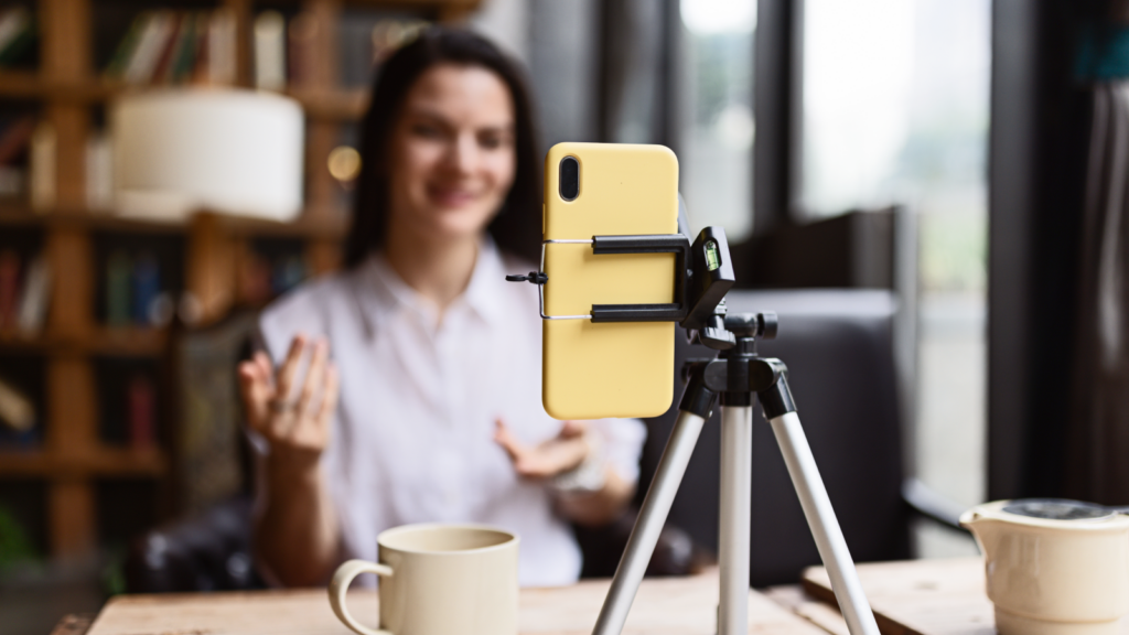 A woman is recording herself with a smartphone, smiling and gesturing while sitting at a table. A coffee cup is on the table.