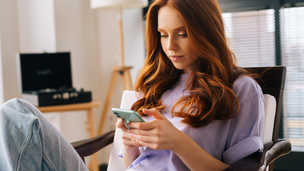 A young woman with long red hair in a lavender top sits comfortably in a chair, focused on her smartphone.