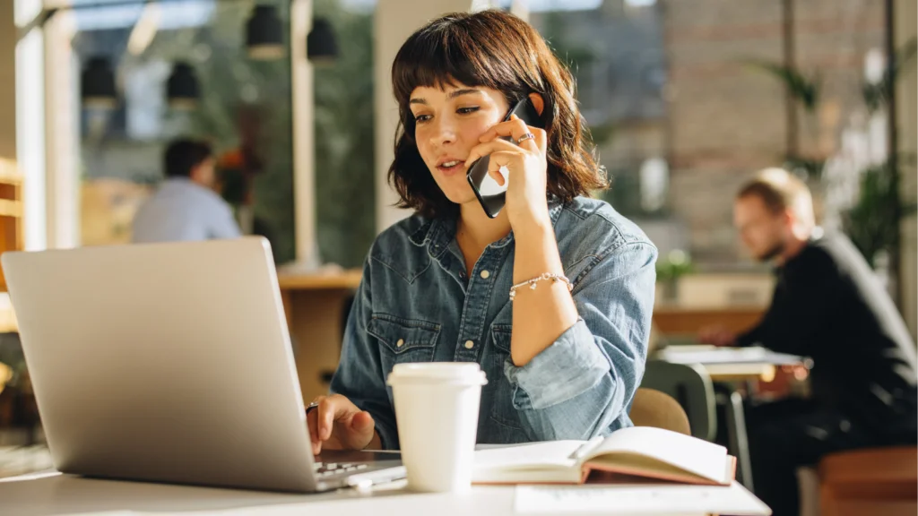 A woman seated at a table, using a laptop and a cell phone, focused on her work.