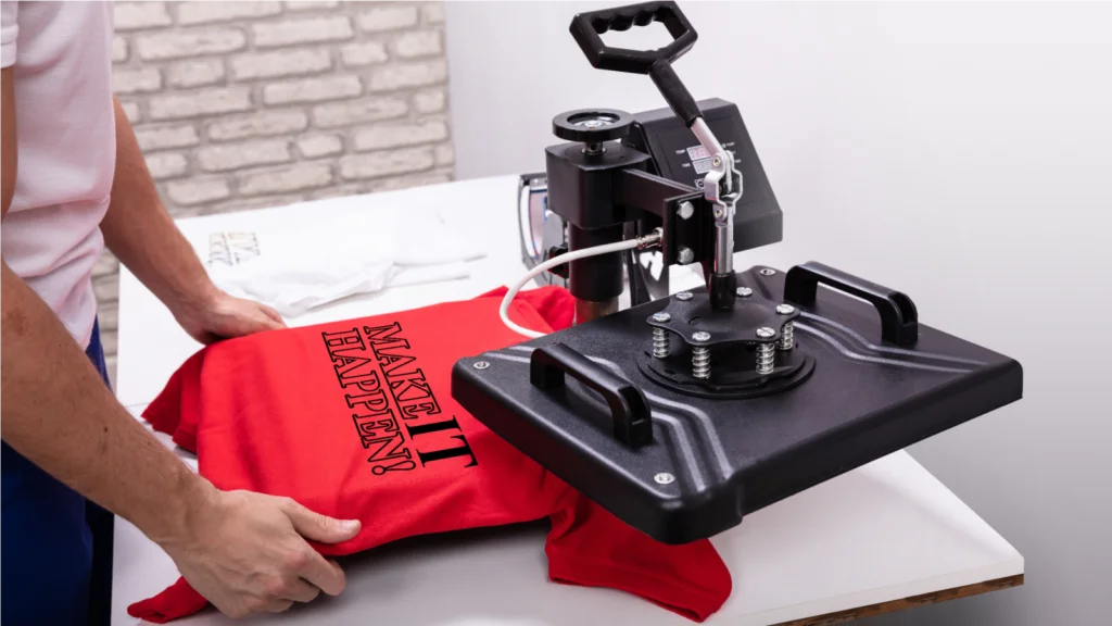 A man operates a machine to print a design on a t-shirt in a workshop setting.