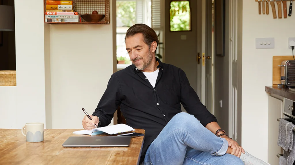 A man in a casual black shirt and jeans is sitting at a kitchen table with a pen and an open notebook, smiling. A laptop and a mug are nearby.