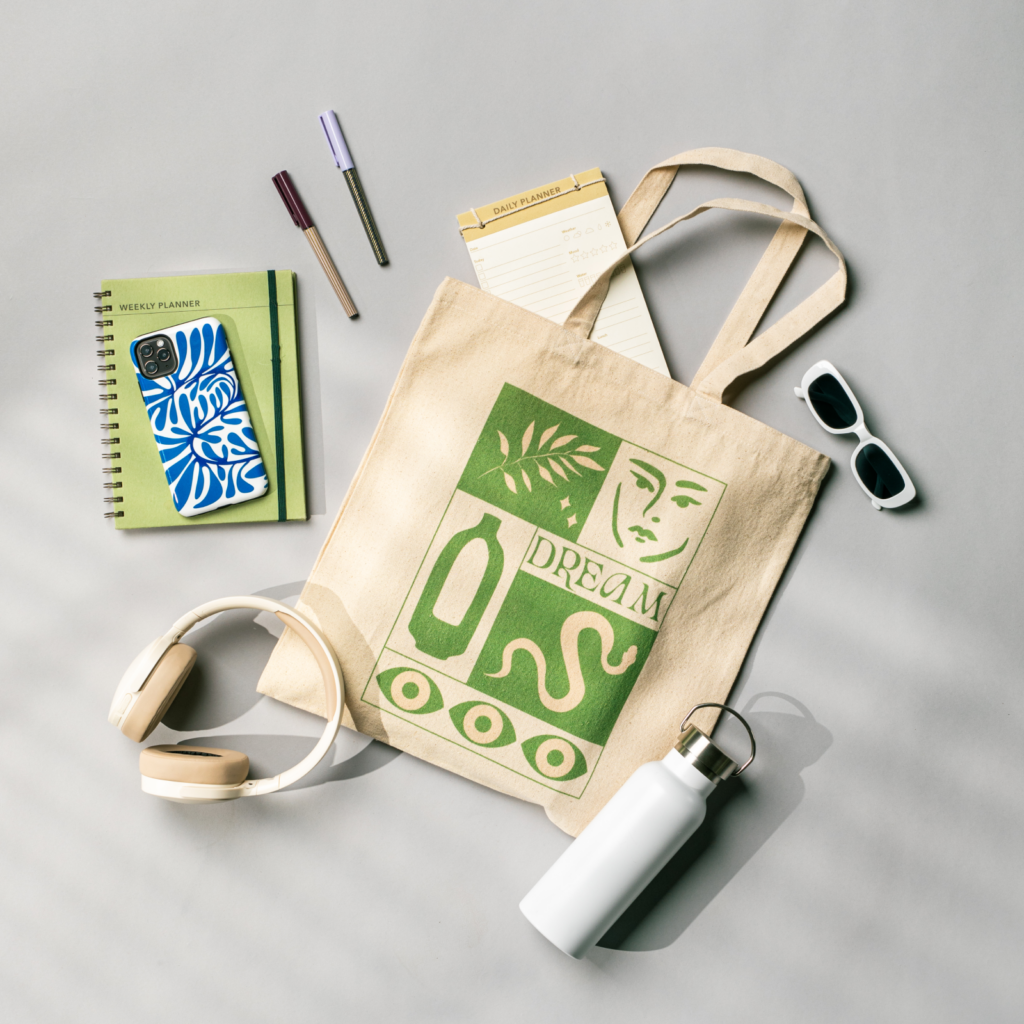Canvas tote bag with green abstract design, “Dream” text, surrounded by a planner, pens, phone case, sunglasses, headphones, and a water bottle.