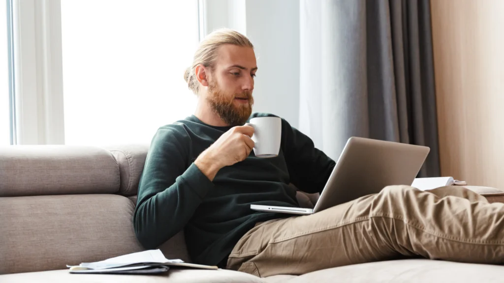 A man with a beard and tied-back hair relaxes on a sofa, sipping from a mug while using a laptop to find the best AI image generators.