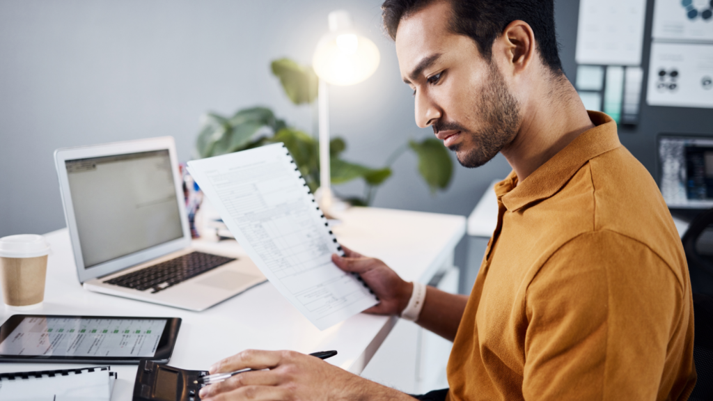 A man in a mustard shirt reviews documents at a desk, using a calculator to calculate sales tax. A laptop, tablet, and cup are nearby.