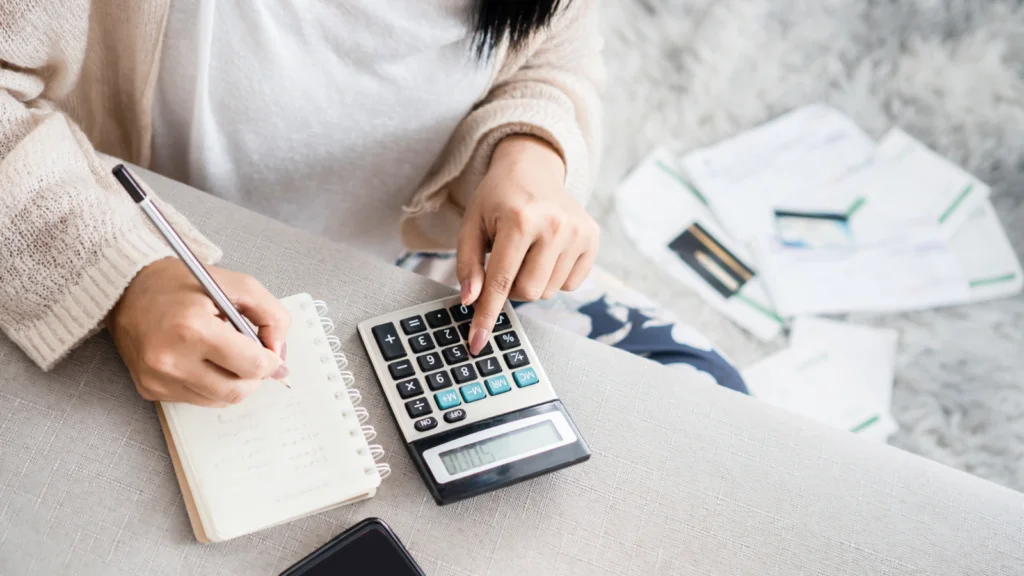 A woman sits on a sofa, using a calculator with one hand and writing in a notebook with the other. Bills and papers are scattered around.