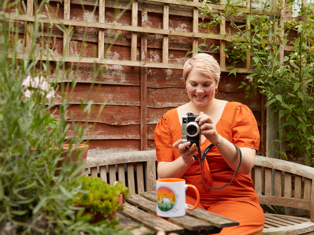 A young woman with short hair is sitting outside, taking a picture of a mug using a camera.