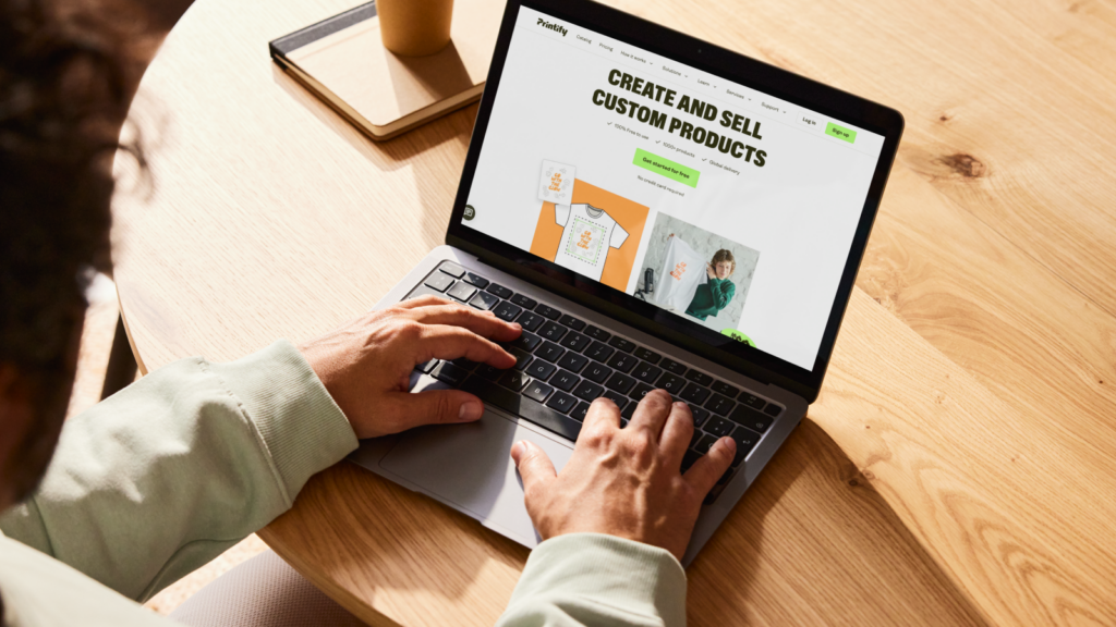 A person is sitting at a table, using a laptop with Printify’s homepage on the screen as an example of how to make money from your phone.