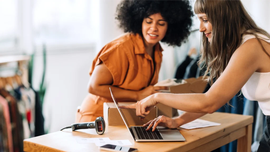 Two women collaborating on a laptop inside a retail store, focused on their work.